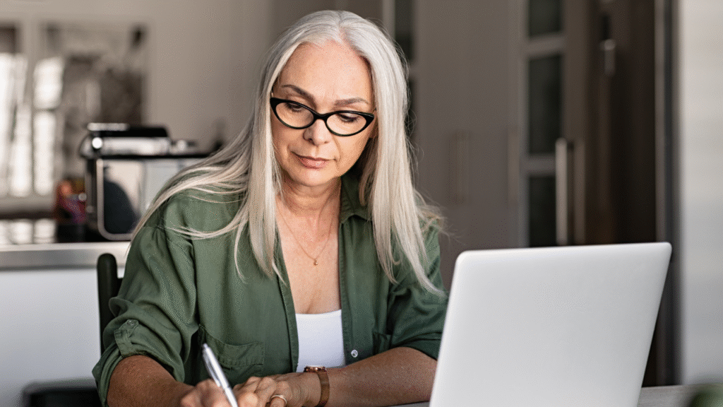 5 Signs You’re Ready to Hire a Transaction Coordinator 1 Woman with gray hair and glasses reviewing paperwork while working on a laptop at a desk, deciding when to hire a transaction coordinator