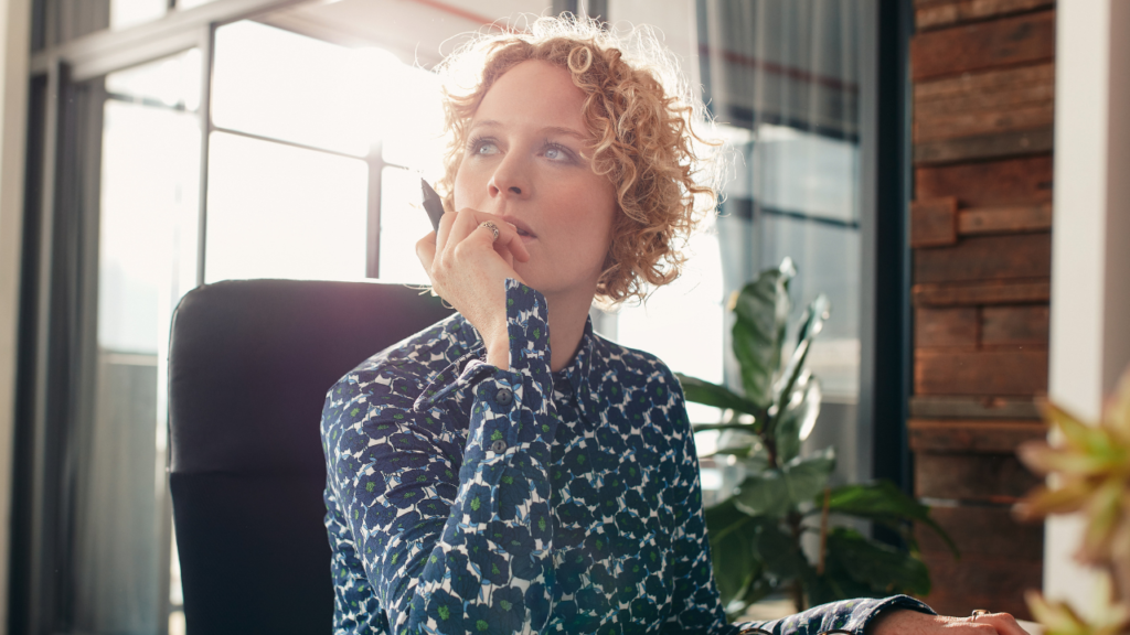 Transaction Coordinator vs. Assistant: What’s the Difference? 1 Curly-haired Realtor sitting thoughtfully at a desk in a modern office, representing the decision between hiring a Transaction Coordinator vs assistant for real estate support.